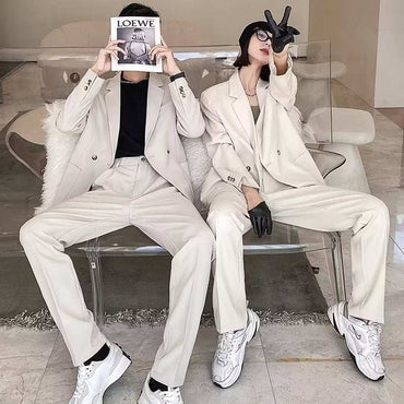 Two models wearing men's casual draping suits in light beige with black inner shirts and white sneakers, seated on a transparent bench in a modern indoor setting
